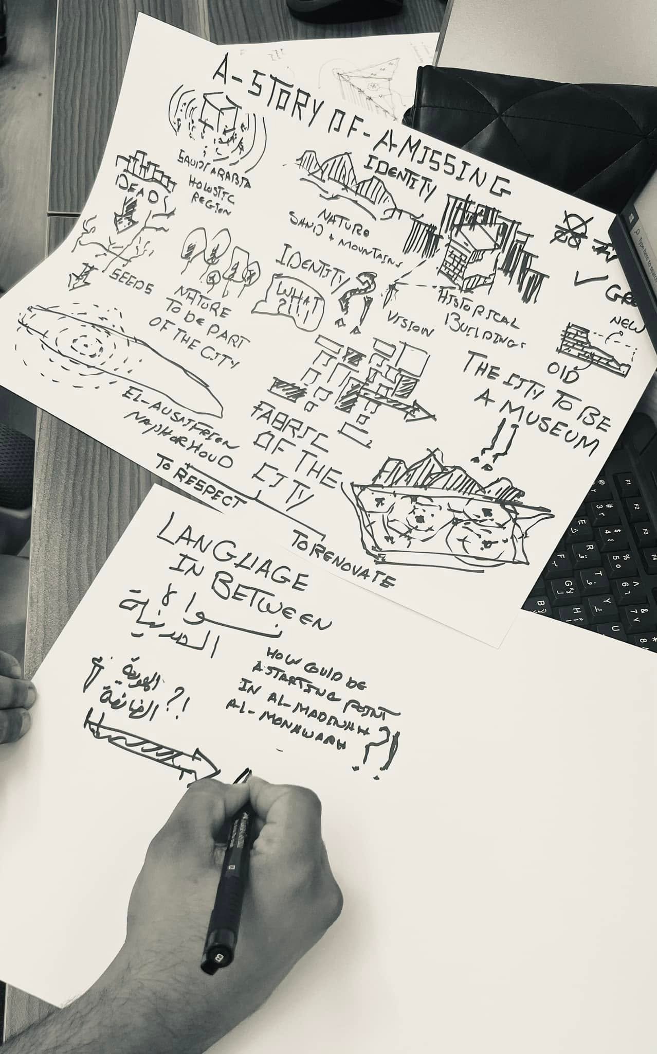 Black-and-white photograph of a hand drawing beside architectural concept sheets about identity, historical buildings, city fabric and a museum.