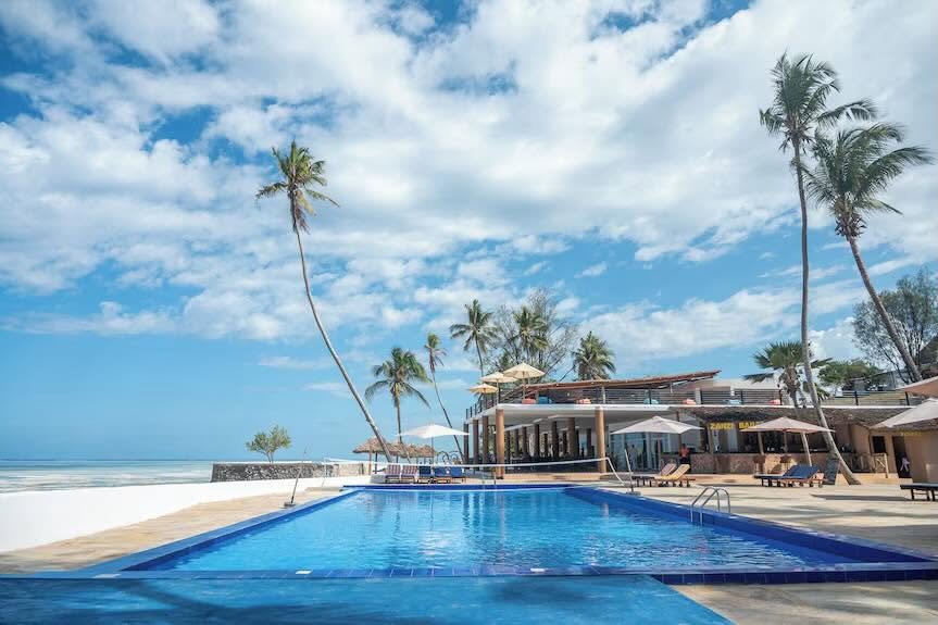 Royal Mandrine Resort site photograph of a blue swimming pool, palms, beachfront building, loungers, and the sea.