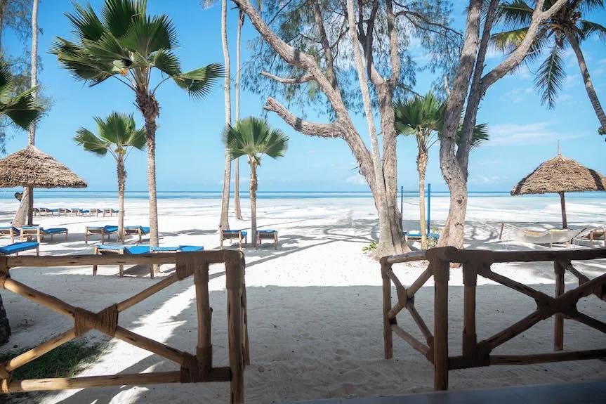 Royal Mandrine Resort site photograph looking from a timber veranda toward a palm-lined beach with loungers and thatched umbrellas.
