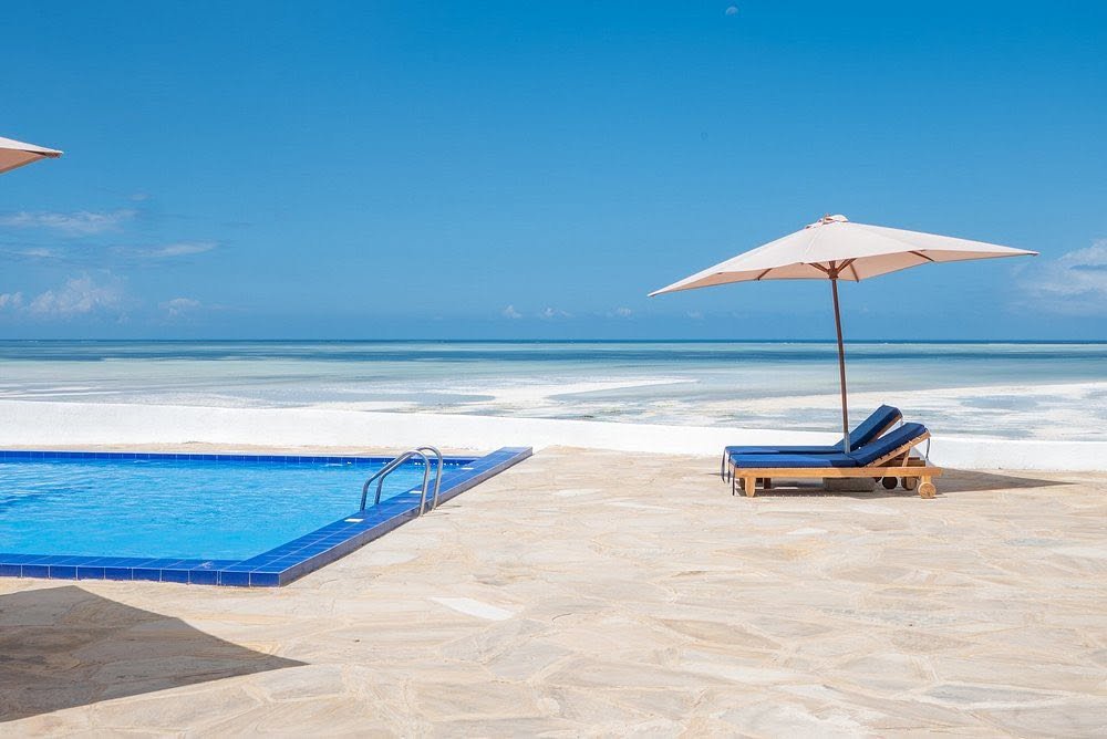 Royal Mandrine Resort site photograph of the pool edge, two blue loungers under an umbrella, and the beach and sea beyond.