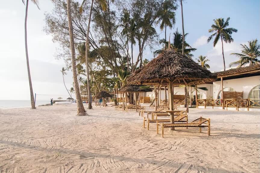 Royal Mandrine Resort beachfront site photograph with thatched shade umbrellas, timber loungers, palms, and white room fronts.