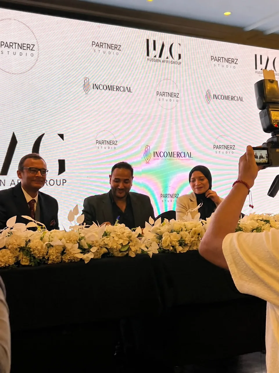 A studio signing moment with three people seated behind a flower-covered table.